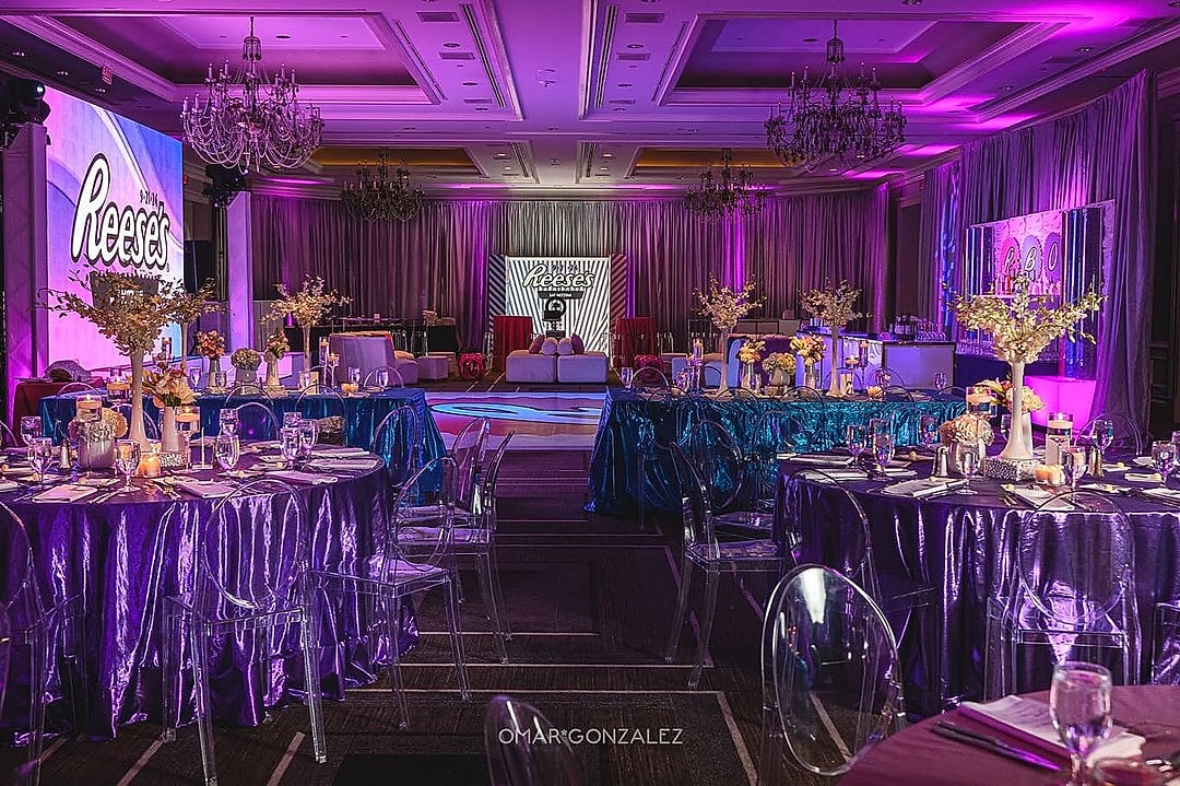 Purple-lit ballroom with tables in shimmering linens and tall centerpieces